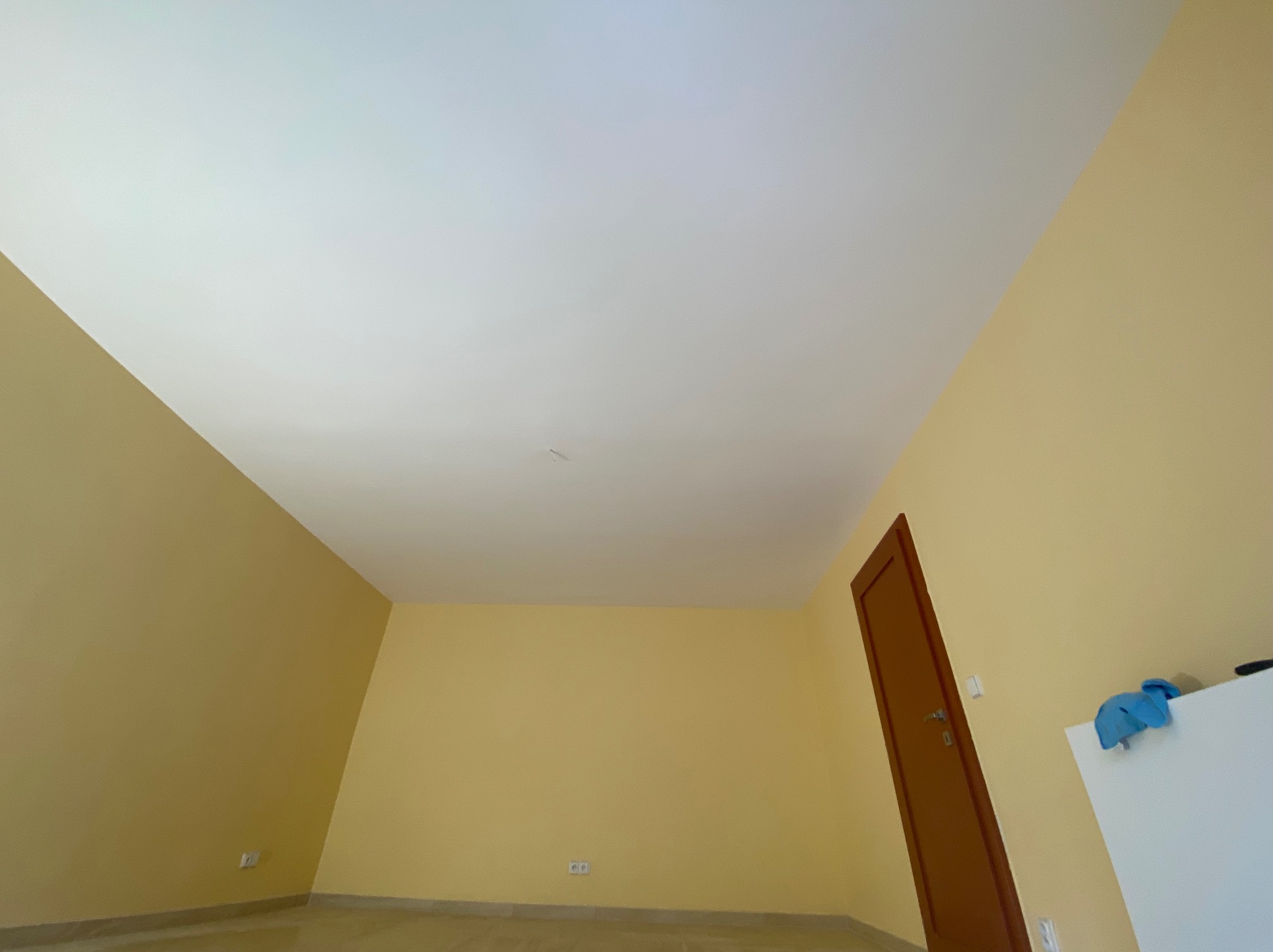 Pomalowane na żółto puste pomieszczenie z białym sufitem, widoczne gniazdka elektryczne, brązowe drzwi i fragment białej szafki z niebieską ścierką.