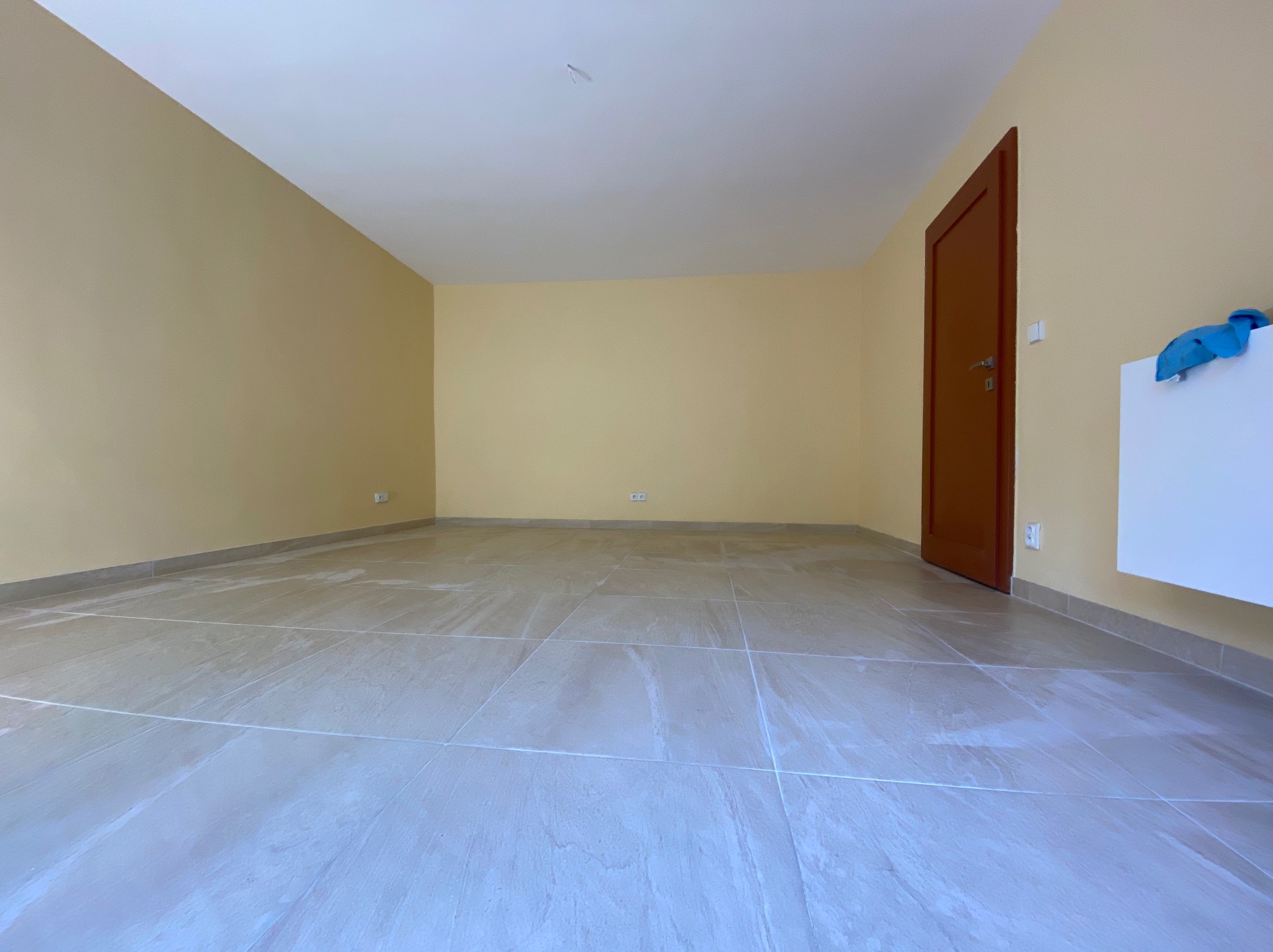 Puste, jasne pomieszczenie po remoncie z beżowymi ścianami, nową, jasną, połyskującą podłogą z płytek imitujących kamień, brązowymi drzwiami po prawej stronie i białym grzejnikiem z niebieską...