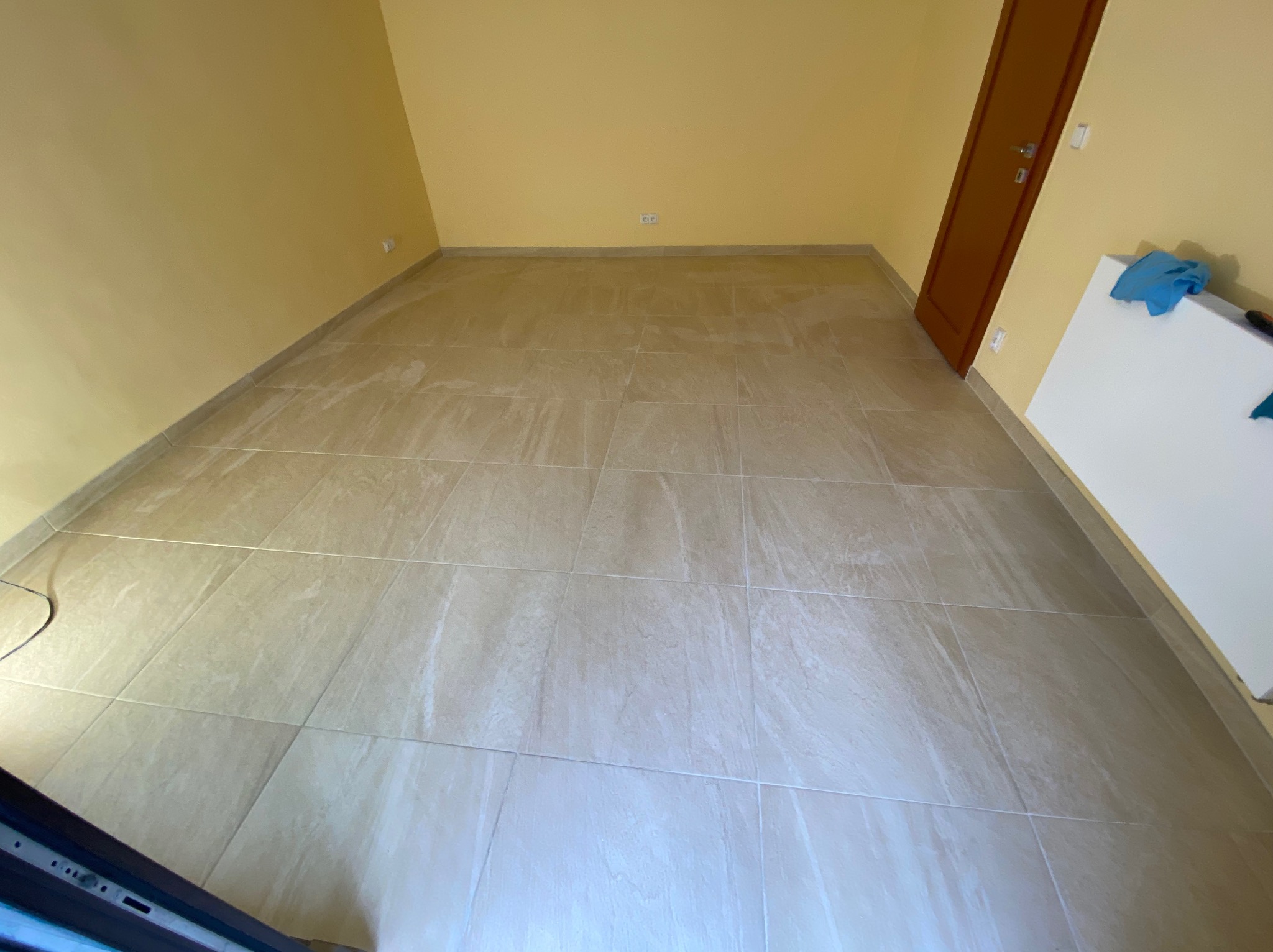 Beżowe płytki podłogowe ułożone w pustym pokoju z żółtymi ścianami i brązowymi drzwiami; widoczny fragment białej płyty z niebieską szmatką.