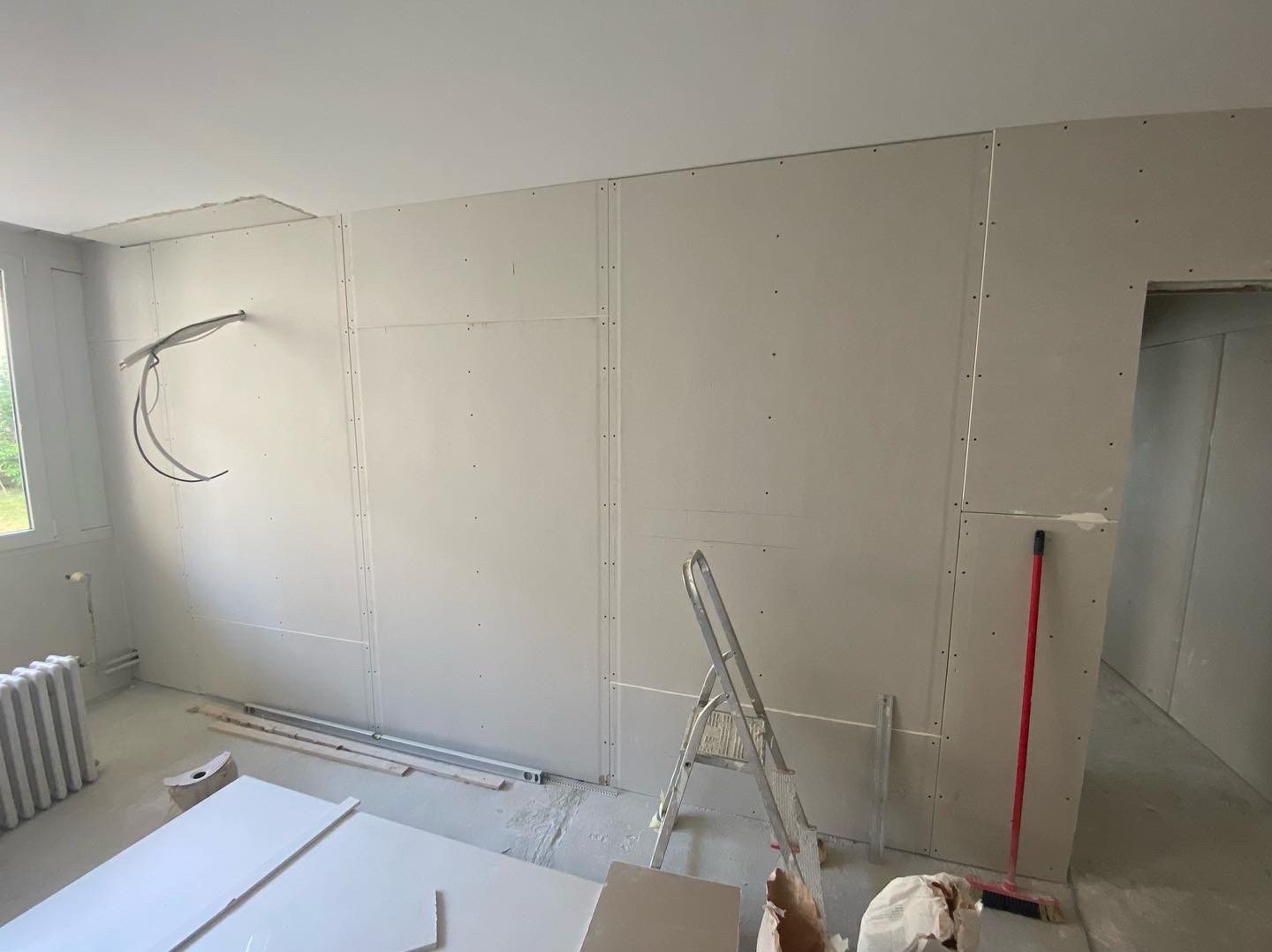 Wnętrze w trakcie montażu płyt kartonowo-gipsowych na ścianach, widoczne narzędzia, drabina aluminiowa i wystające przewody elektryczne.