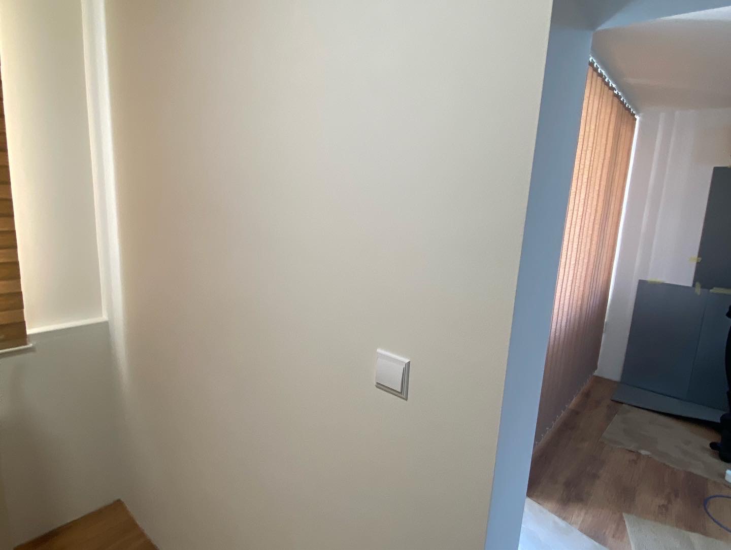 Świeżo pomalowana ściana w jasnym odcieniu z zamontowanym włącznikiem światła, widoczne rolety pionowe i fragment podłogi.
