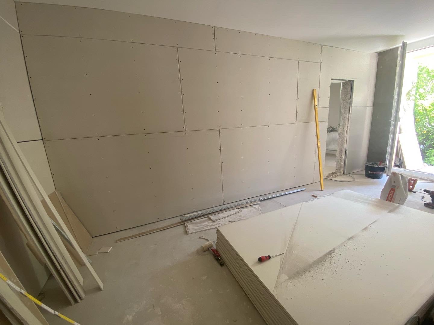 Montaż płyt kartonowo-gipsowych na ścianie z widocznymi wkrętami, poziomica oparta o ścianę, stos płyt na podłodze, widok na otwór drzwiowy i okno w tle.