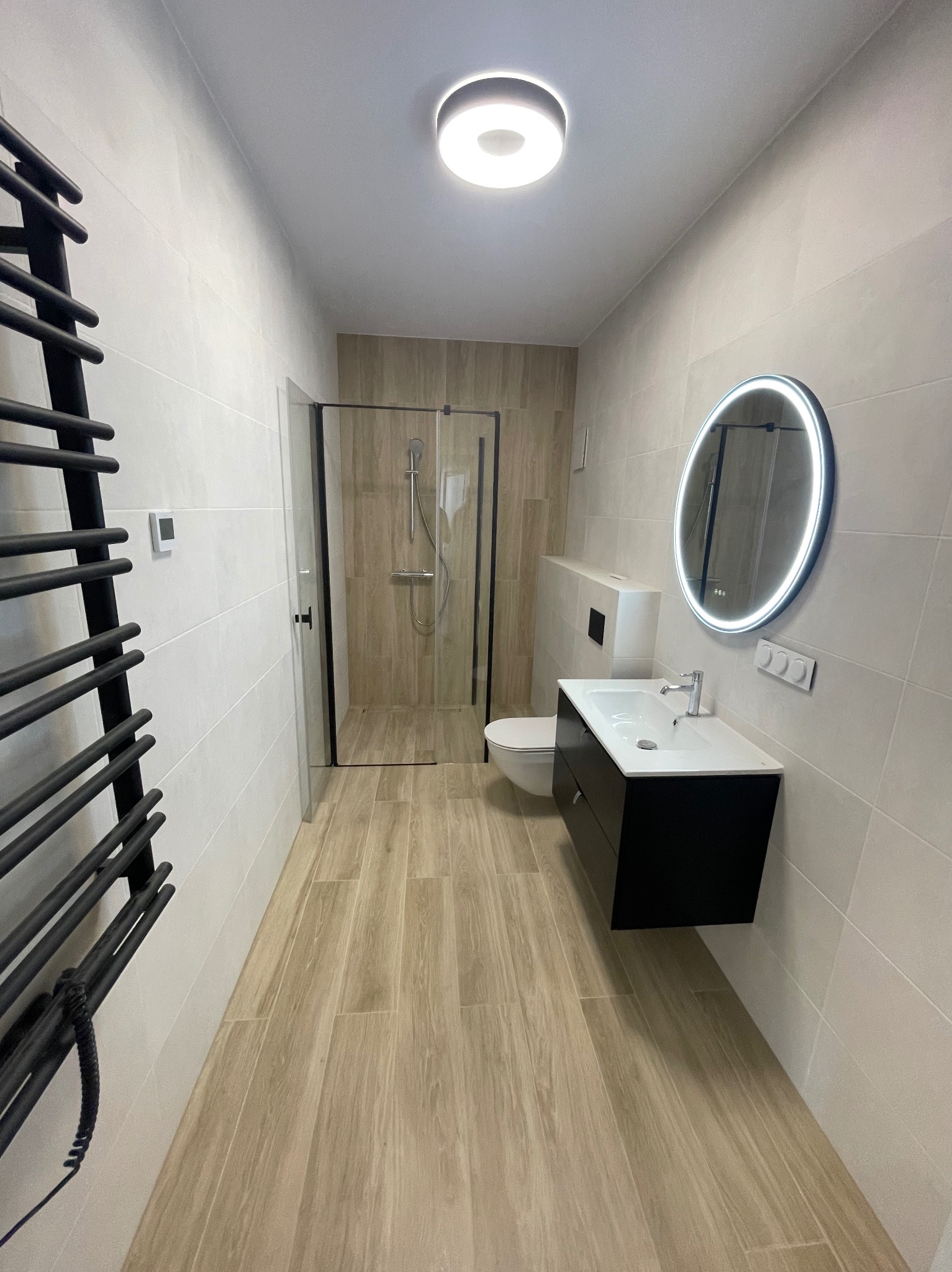 Wnętrze wykończonej łazienki w jasnych barwach z kabiną prysznicową, umywalką z szafką i okrągłym lustrem z podświetleniem LED oraz grzejnikiem drabinkowym.