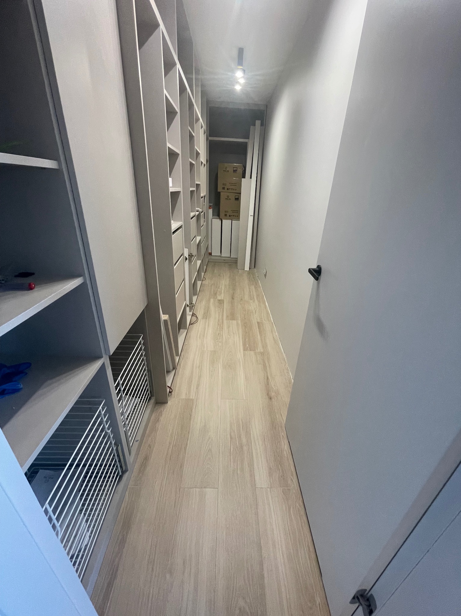 Wąskie przejście z zamontowaną, szarą zabudową szaf z półkami i szufladami po lewej stronie. Na podłodze panele imitujące drewno. Ściany pomalowane na szaro.