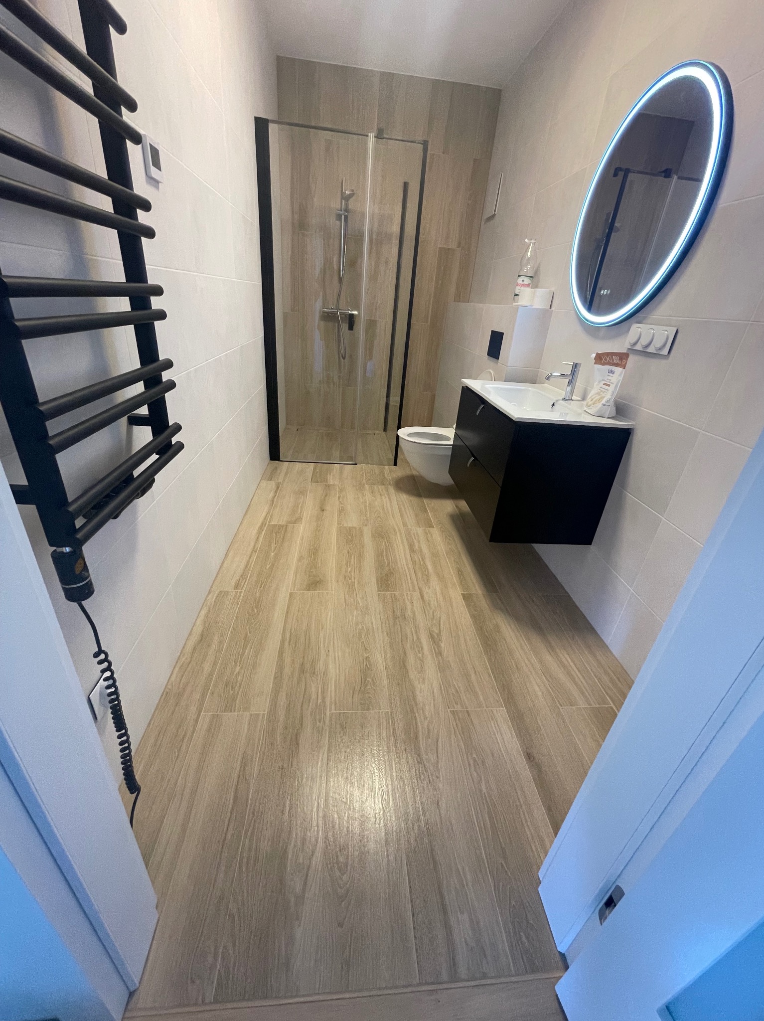 Nowoczesna łazienka po remoncie: kabina prysznicowa, umywalka z szafką, podświetlane lustro i grzejnik drabinkowy. Płytki imitujące drewno na podłodze i ścianach.