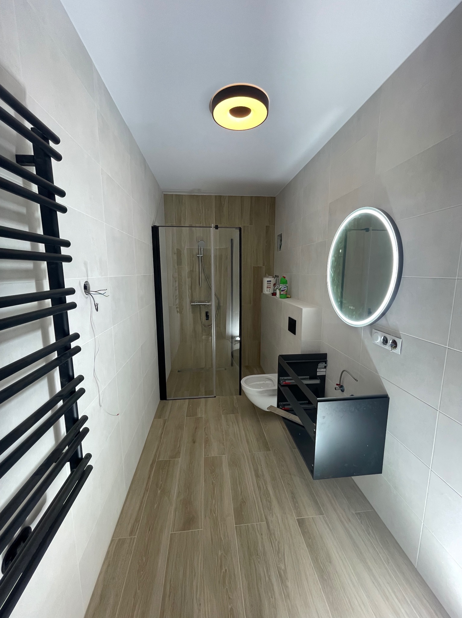 Nowoczesna łazienka po remoncie: kabina prysznicowa z czarną ramą, podświetlane lustro nad umywalką bez szafki i grzejnik drabinkowy na tle beżowych płytek.