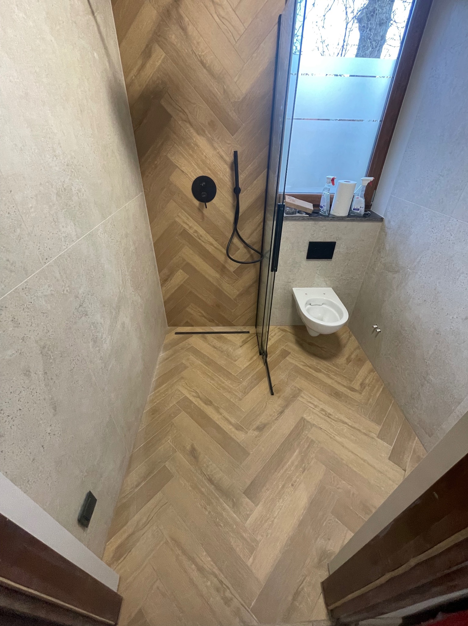 Nowoczesna łazienka z kabiną prysznicową typu walk-in, wykończona płytkami w odcieniach beżu ułożonymi w jodełkę. Widoczna toaleta pod oknem i czarna armatura.