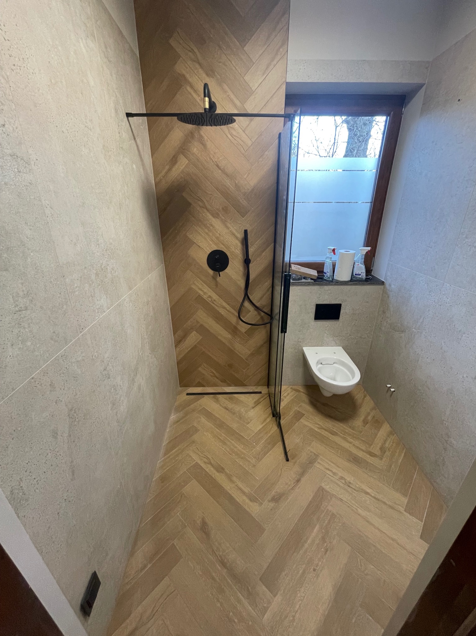 Nowoczesna łazienka z kabiną prysznicową typu walk-in. Jasne płytki imitujące drewno ułożone w jodełkę na podłodze i ścianie prysznica. Minimalistyczna armatura w czarnym macie.