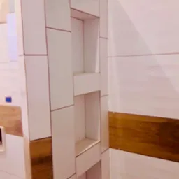 Wnęki w ścianie wyłożonej białymi i drewnopodobnymi płytkami w trakcie montażu, widoczne niedociągnięcia w górnej części ściany.