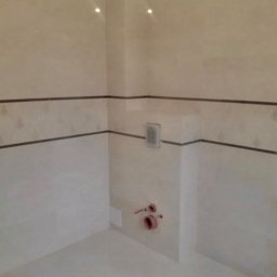 Krzysztof Lizner - Wykończona łazienka z beżowymi i białymi płytkami, widoczne rury instalacji wodnej i odpływ, dekoracyjny pasek płytek na ścianie.