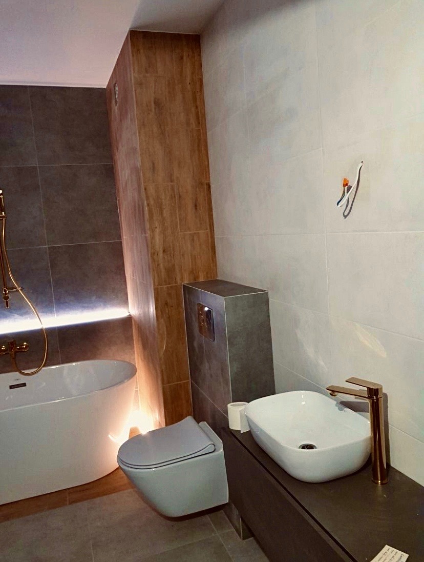 Nowoczesna łazienka z podświetlaną wanną, wiszącą toaletą i umywalką nablatową z armaturą w kolorze różowego złota, wykończona płytkami w odcieniach szarości i beżu, z widocznymi przewodami...