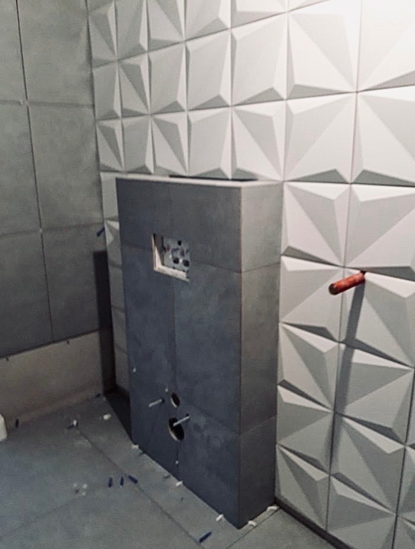 Montaż podtynkowej spłuczki WC obłożonej szarymi płytkami, widoczne wystające rurki instalacyjne, tło z białych, trójwymiarowych paneli ściennych.