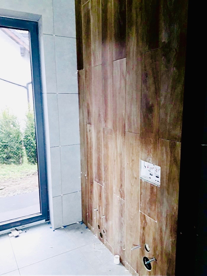 Ściana w łazience w trakcie układania płytek imitujących drewno, widoczne otwory na instalacje i częściowo ułożone białe płytki na sąsiedniej ścianie i podłodze, okno z widokiem na zieleń na zewnątrz.