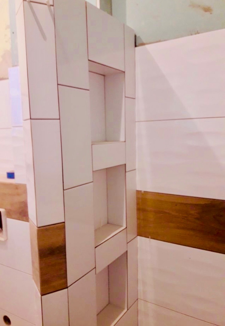 Wnęki w ścianie wyłożonej białymi i drewnopodobnymi płytkami w trakcie montażu, widoczne niedociągnięcia w górnej części ściany.