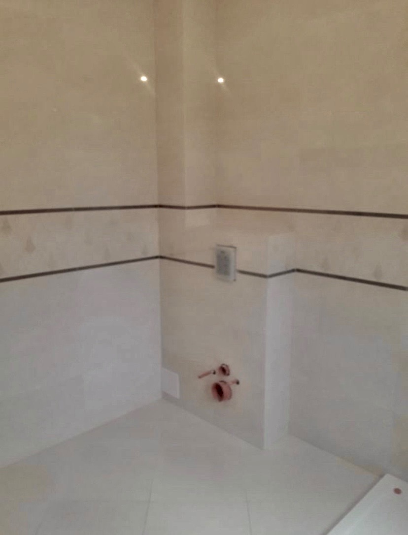 Wykończona łazienka z beżowymi i białymi płytkami, widoczne rury instalacji wodnej i odpływ, dekoracyjny pasek płytek na ścianie.