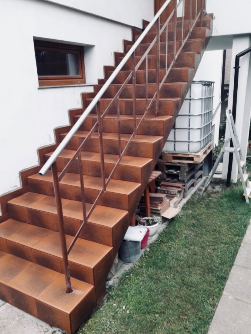 Zewnętrzne schody wyłożone brązowymi płytkami, z metalową balustradą, prowadzące do wejścia domu z małym oknem; w tle biały zbiornik na paletach i drabina.