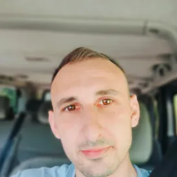 Mężczyzna w niebieskiej koszulce i pasach bezpieczeństwa, uśmiechający się, z widocznym wnętrzem samochodu w tle.