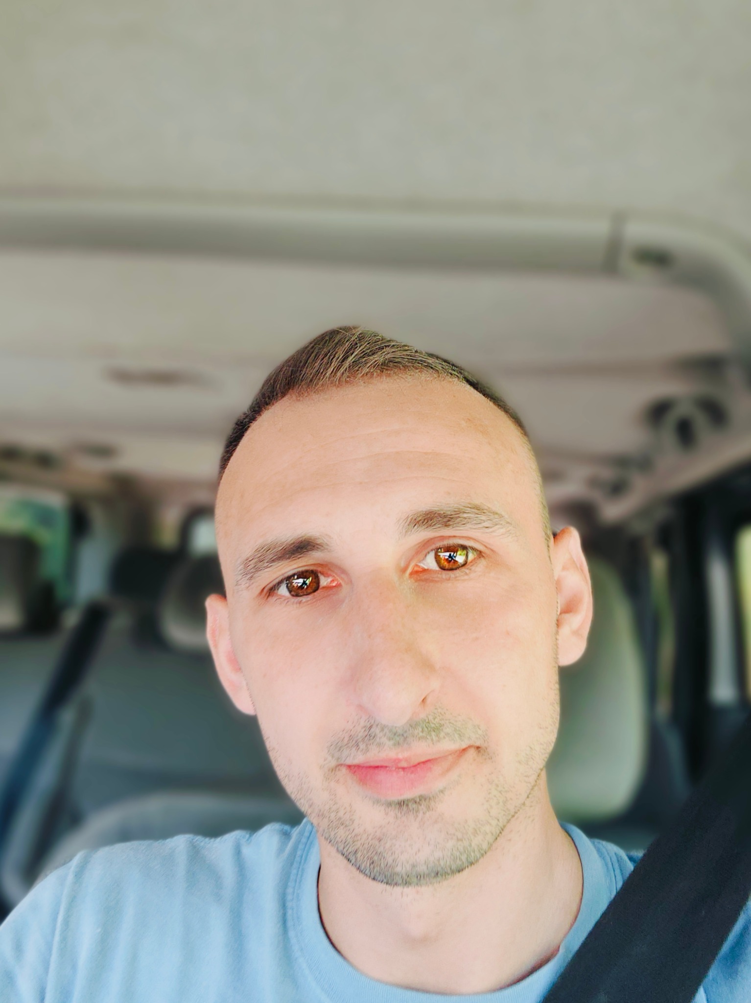 Mężczyzna w niebieskiej koszulce i pasach bezpieczeństwa, uśmiechający się, z widocznym wnętrzem samochodu w tle.