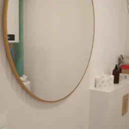 Okrągłe lustro w złotej ramie zawieszone na białej ścianie w łazience, widoczne zawory, pudełko z chusteczkami i dozownik mydła na blacie.