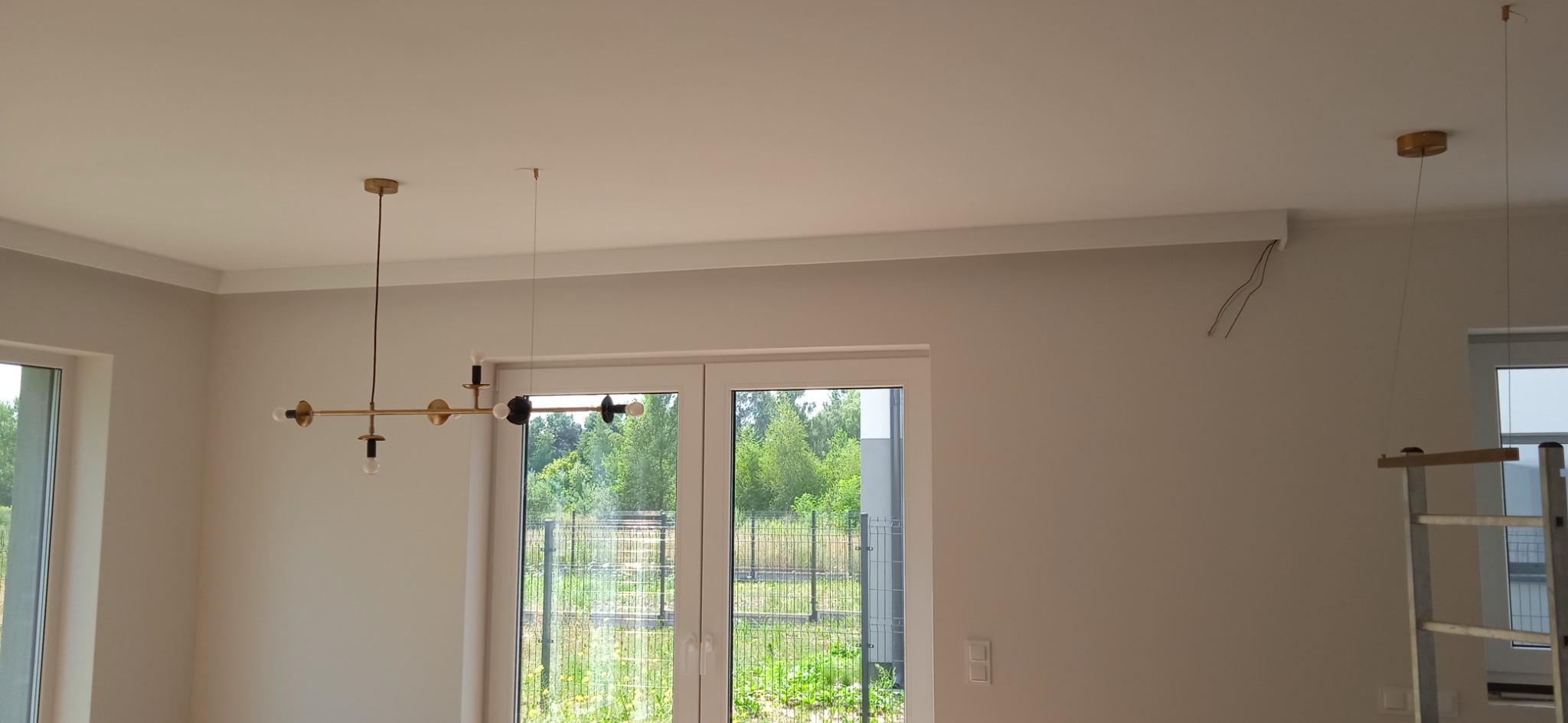 Nowoczesny, złoty żyrandol z czarnymi elementami w jasnym wnętrzu w trakcie wykańczania, widoczne wystające kable i drabina.