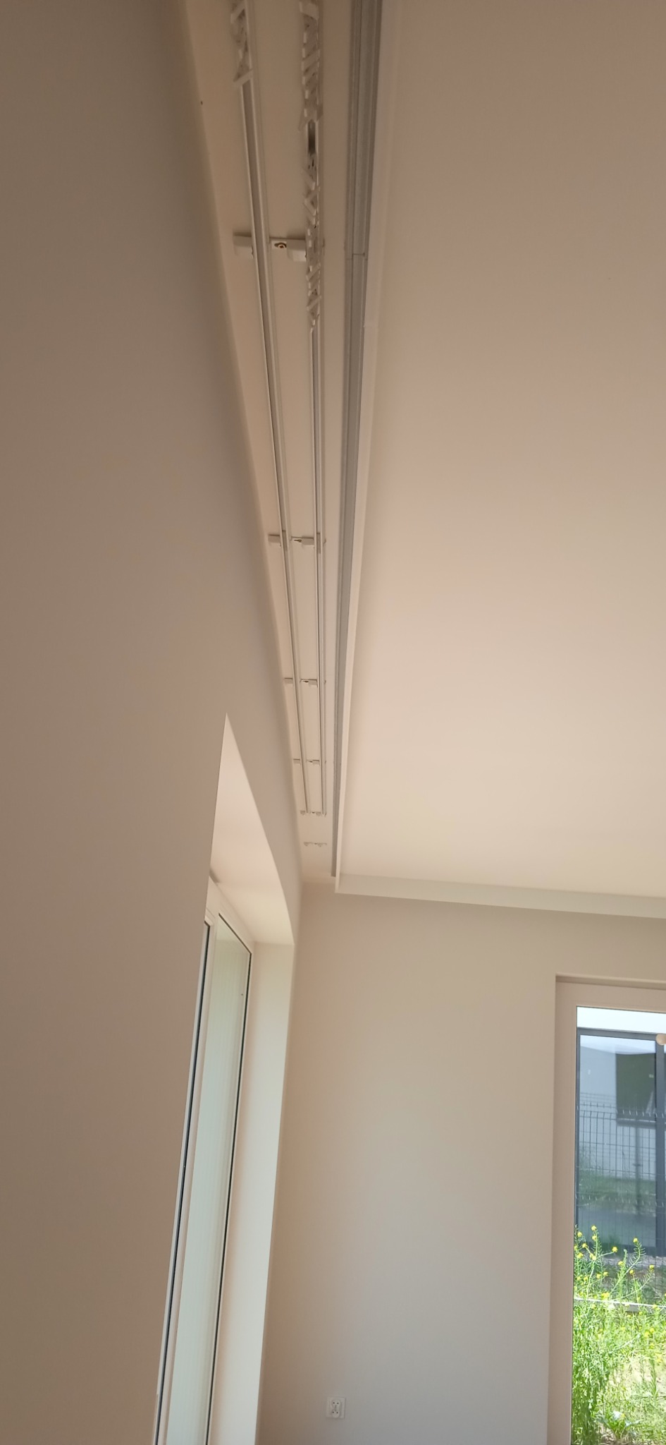 Zamontowane szyny sufitowe karnisza w jasnym pokoju, widoczne okna i fragment roślinności za oknem.