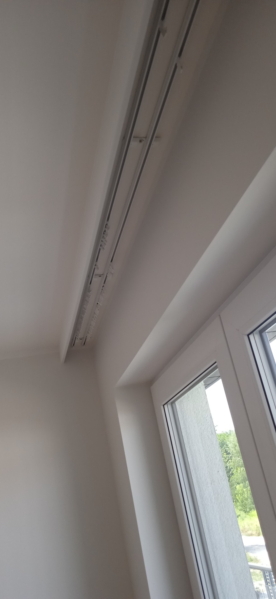 Zamontowane podwójne szyny sufitowe karnisza z zaczepami, widoczne na tle białego sufitu i okna z widokiem na zieleń.