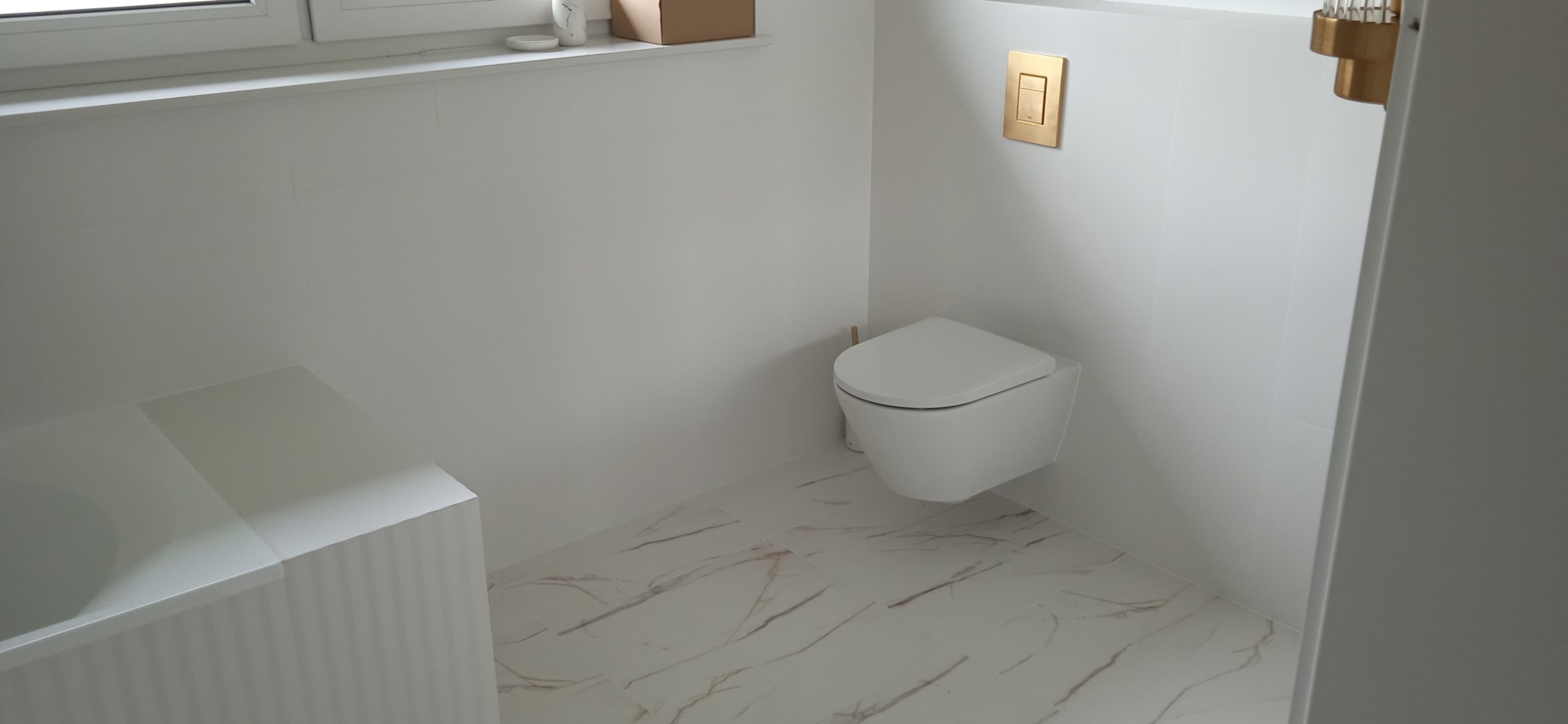 Nowoczesna, minimalistyczna toaleta podwieszana w białej łazience z marmurową podłogą i złotym przyciskiem spłuczki.