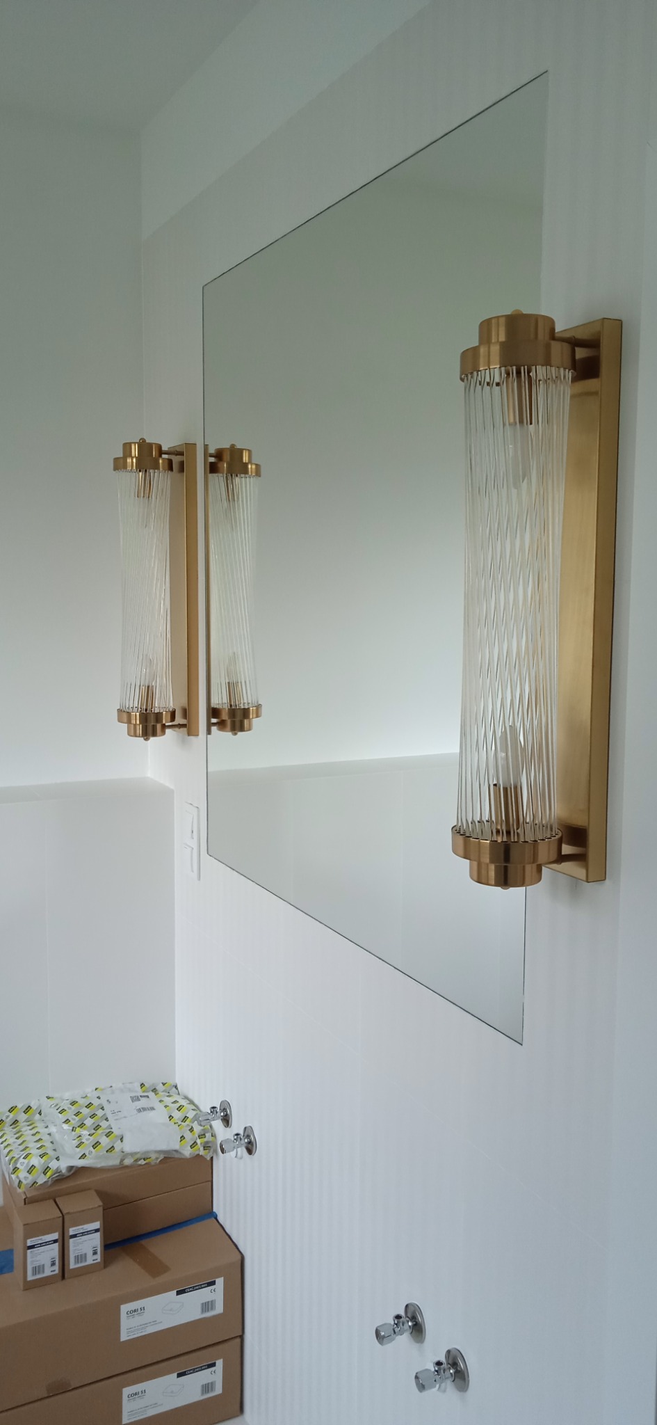 Zamontowane, złote kinkiety z przezroczystymi, żłobionymi kloszami po obu stronach lustra w białej łazience, na dole kartony z armaturą.