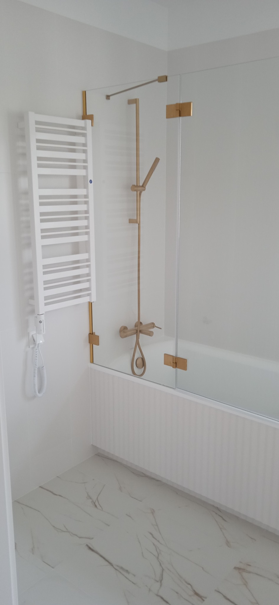 Nowoczesna łazienka w bieli z akcentami złotego koloru: grzejnik drabinkowy, bateria prysznicowa i szklany parawan nawannowy z zawiasami.