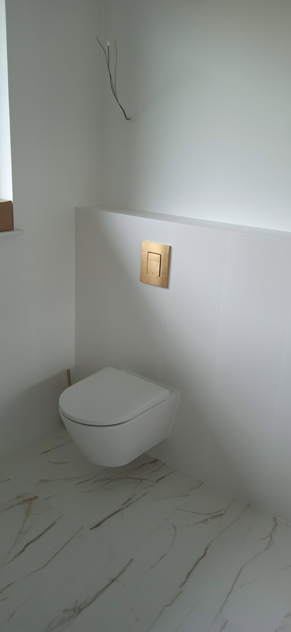 Podwieszana toaleta w nowoczesnej łazience z białymi płytkami imitującymi marmur na podłodze i ścianie, złoty przycisk spłuczki, widoczne wystające przewody elektryczne.
