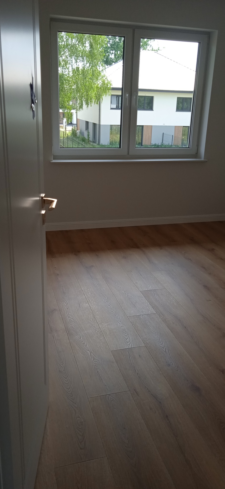 Wnętrze pokoju z nową podłogą z paneli drewnopodobnych, widok na okno z widokiem na dom i zieleń, częściowo otwarte białe drzwi z złotą klamką po lewej.
