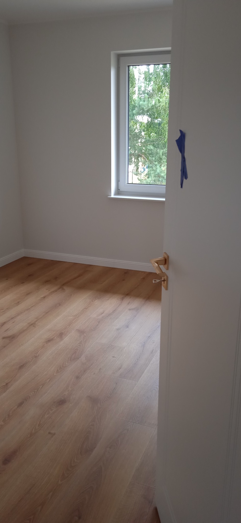 Wykończone wnętrze pokoju w Podolszynie Nowym: panele podłogowe w odcieniach drewna, białe ściany, okno z widokiem na zieleń i częściowo otwarte białe drzwi z złotą klamką.