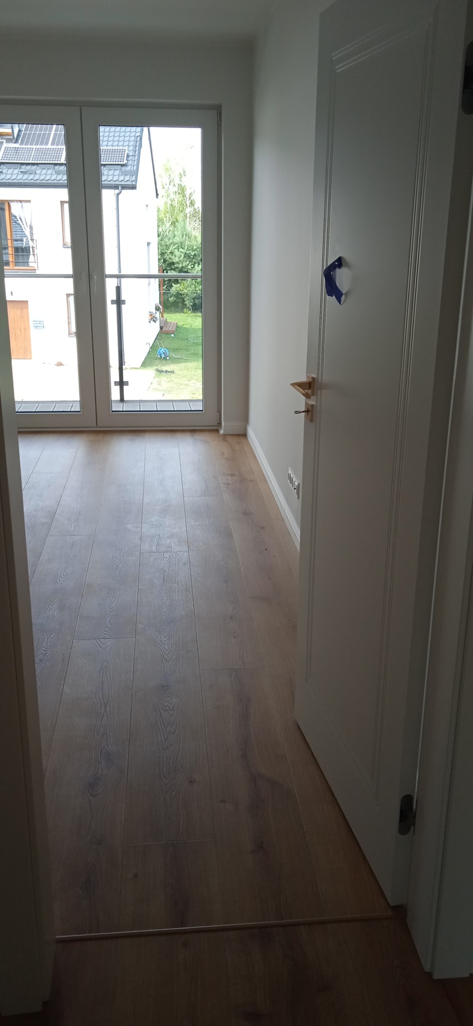 Wykończone wnętrze: parkiet ułożony w jasnym pokoju z widokiem na ogród przez przeszklone drzwi balkonowe, białe ściany i drzwi z mosiężną klamką, zabezpieczone taśmą malarską.