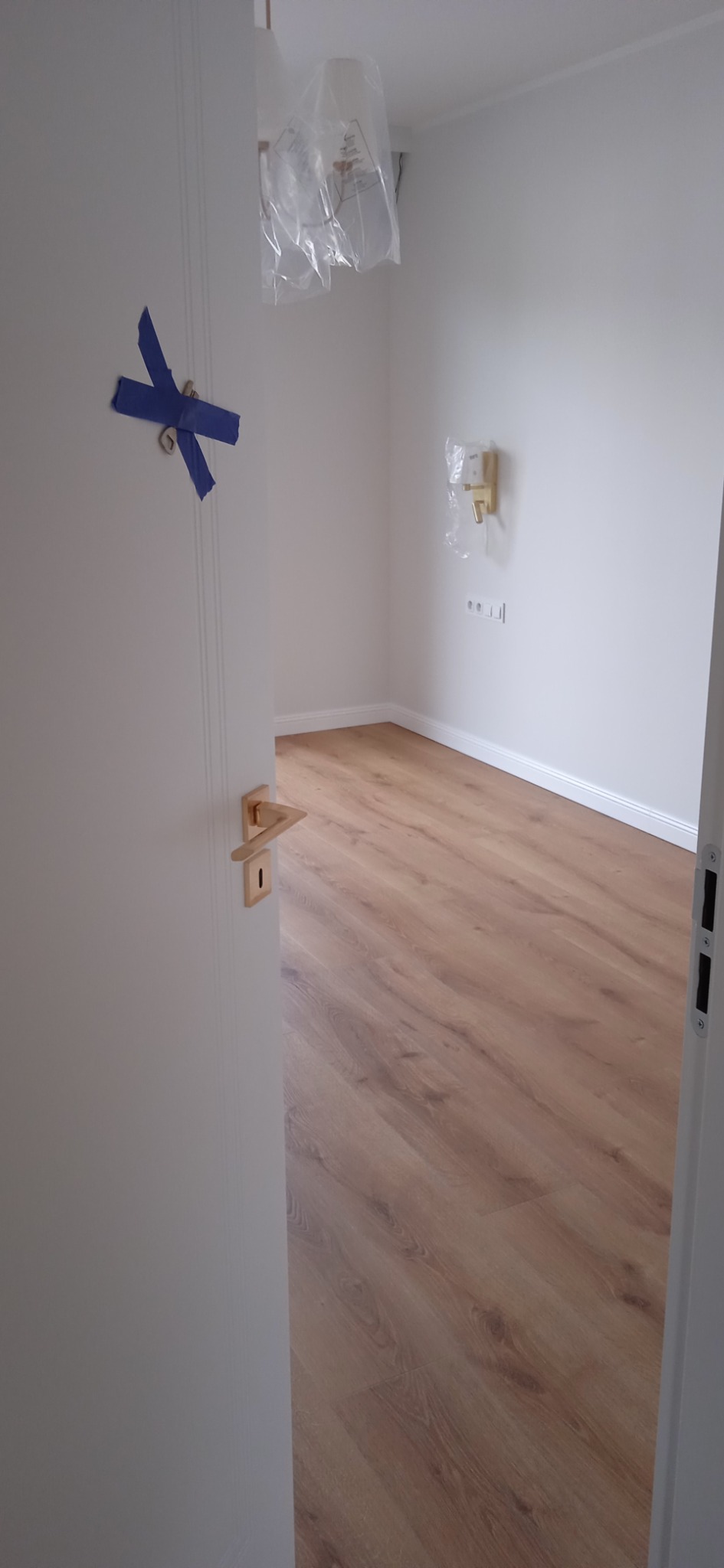 Wykończony pokój z drewnianą podłogą, widok przez uchylone białe drzwi z niebieską taśmą zabezpieczającą, osprzęt elektryczny na ścianie