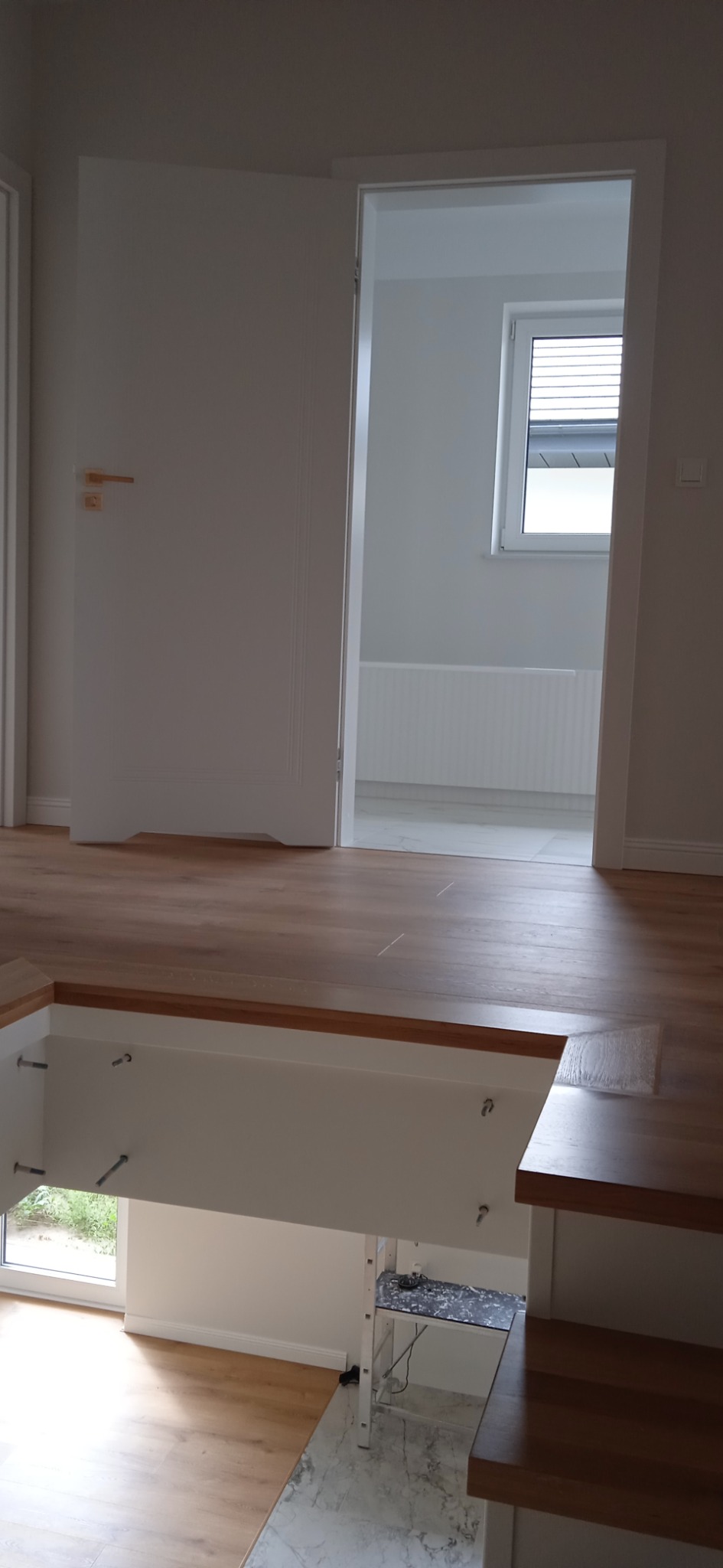 Wnętrze mieszkania w trakcie prac wykończeniowych, widoczne schody z drewnianymi stopniami, otwarta biała stolarka drzwiowa i fragment pomieszczenia z grzejnikiem pod oknem.