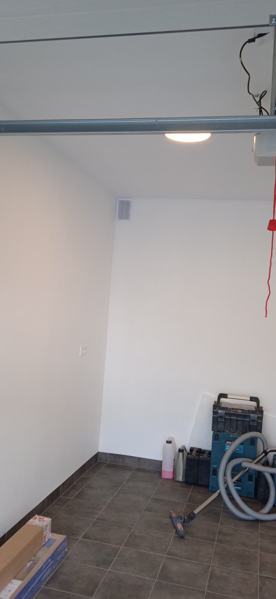 Pusty garaż z szarą podłogą z płytek, białe ściany z kratką wentylacyjną, narzędzia i środki czystości w rogu, przygotowany do prac wykończeniowych.