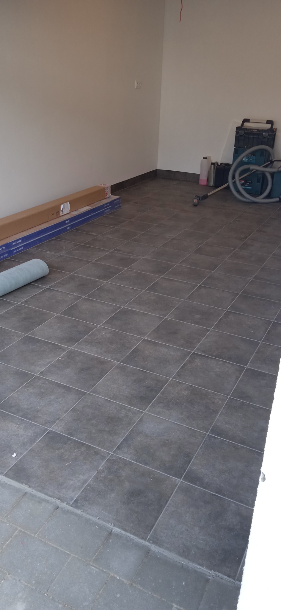 Ułożone szare płytki podłogowe w pomieszczeniu, z widocznymi materiałami budowlanymi i odkurzaczem, sugerujące prace wykończeniowe.