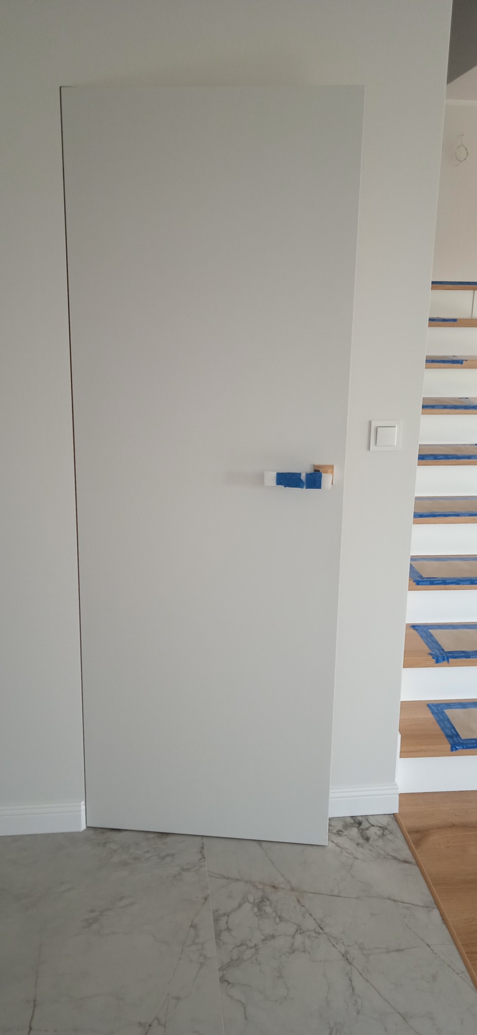 Białe, ukryte drzwi z maskowaną klamką zabezpieczoną niebieską taśmą, widoczne białe ściany, marmurowe płytki podłogowe i schody z drewnianymi stopniami zabezpieczonymi folią.