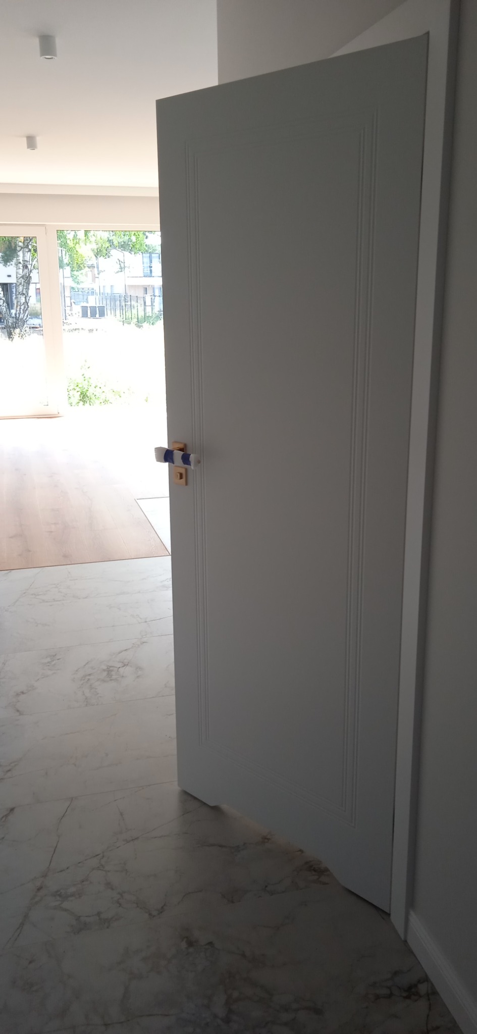 Otwarta biała, płycinowa drzwi wewnętrzne z osłoniętą klamką, widoczna marmurowa posadzka i parkiet w tle.