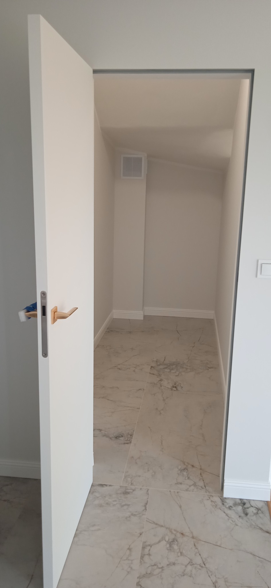 Widok na małą garderobę z marmurową podłogą przez uchylone białe drzwi z drewnianą klamką.