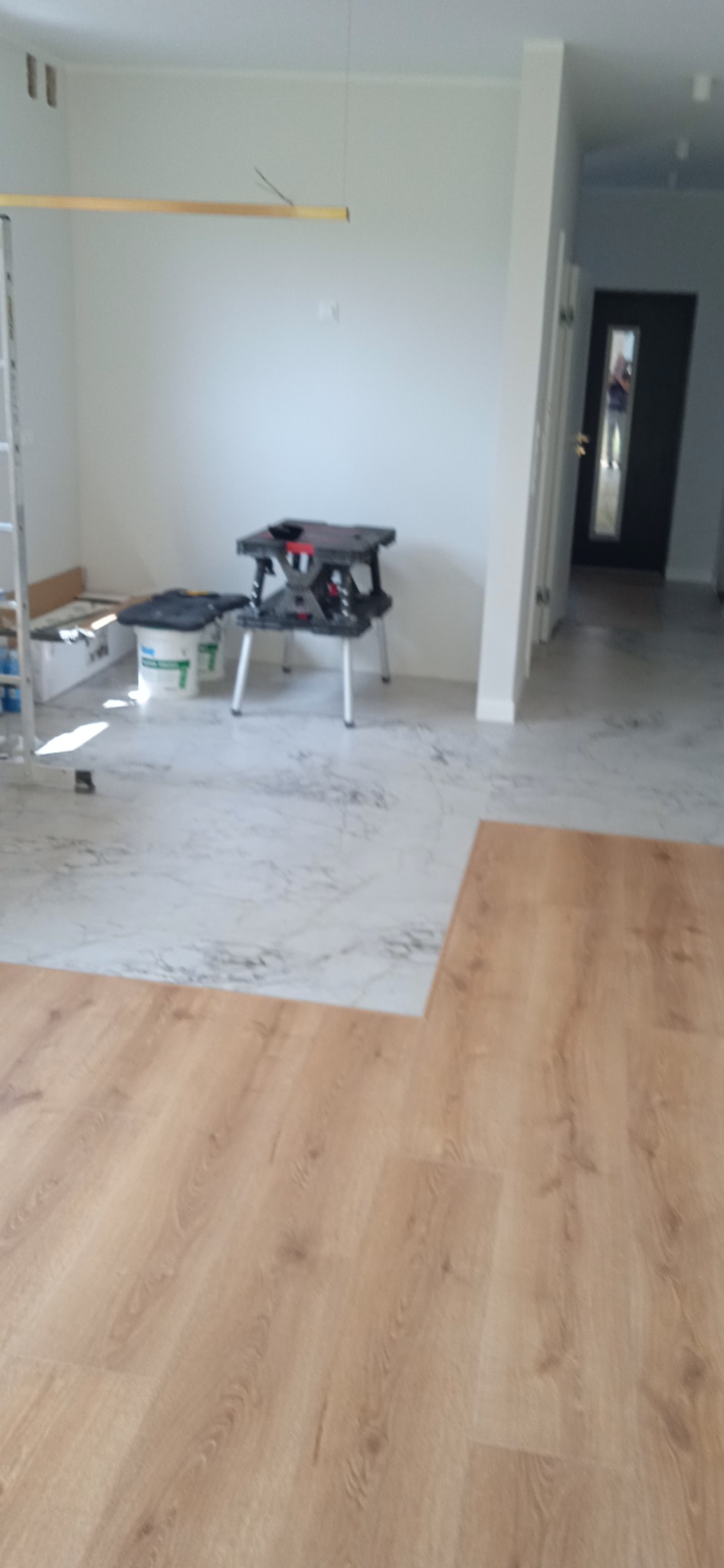 Fragment podłogi w trakcie wymiany: położone panele laminowane w odcieniach brązu sąsiadują z fragmentem podłogi wyłożonym płytkami imitującymi marmur. W tle widoczny stół warsztatowy, drabina...