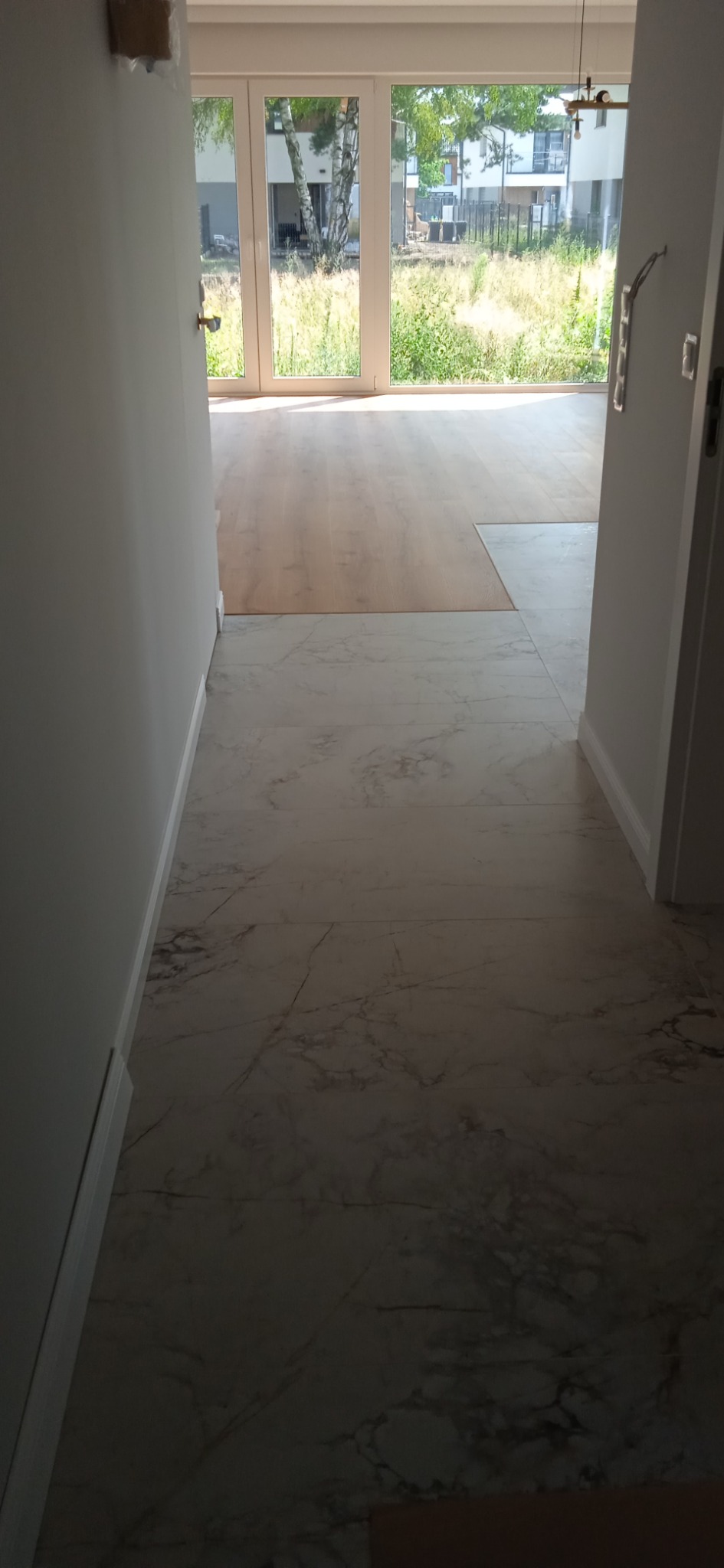 Wykończona podłoga z białego marmuru w przejściu do jasnego salonu z drewnianą podłogą i dużymi oknami z widokiem na zieleń.