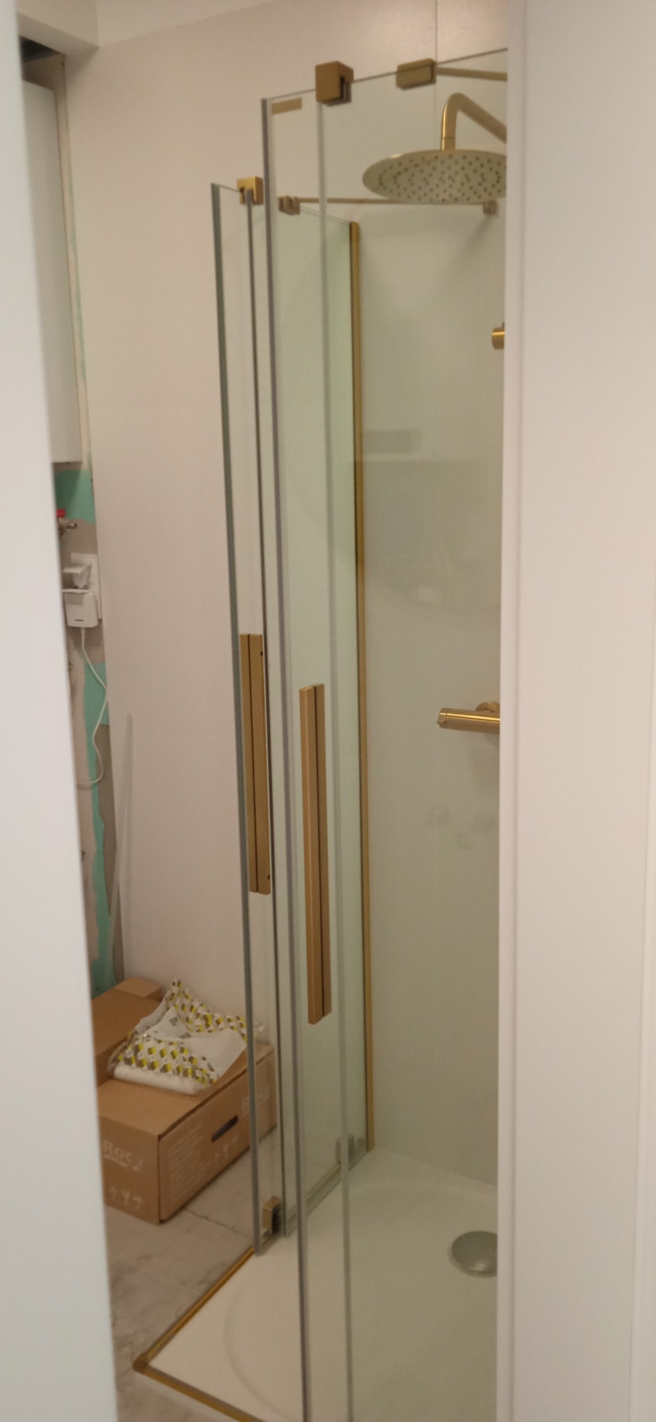 Kabina prysznicowa z transparentnego szkła z złotymi okuciami i uchwytami, widoczna w trakcie montażu, obok kartonowe pudła i ślady remontu na ścianie.