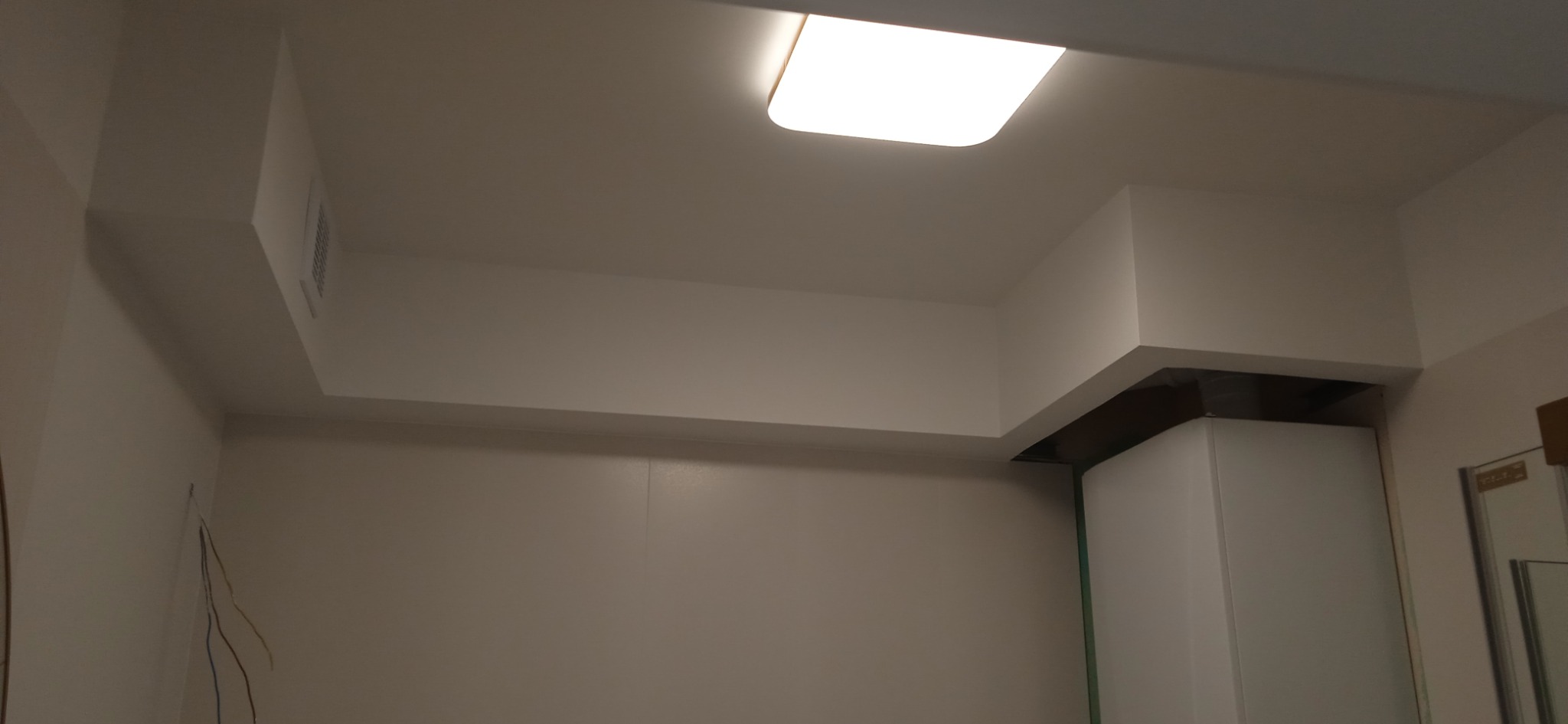 Wykończony sufit podwieszany z wbudowanym oświetleniem LED w pomieszczeniu z pomalowanymi ścianami na jasny odcień beżu, widoczne wystające przewody elektryczne na ścianie.