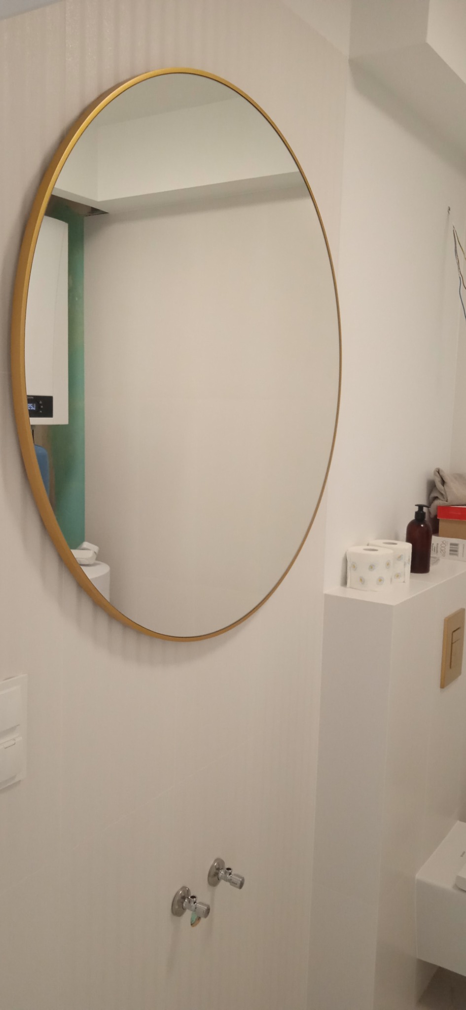 Okrągłe lustro w złotej ramie zawieszone na białej ścianie w łazience, widoczne zawory, pudełko z chusteczkami i dozownik mydła na blacie.