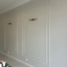 Ściana w jasnych odcieniach szarości z białymi, dekoracyjnymi panelami ściennymi i złotymi kinkietami, widoczne narzędzia malarskie na stole.