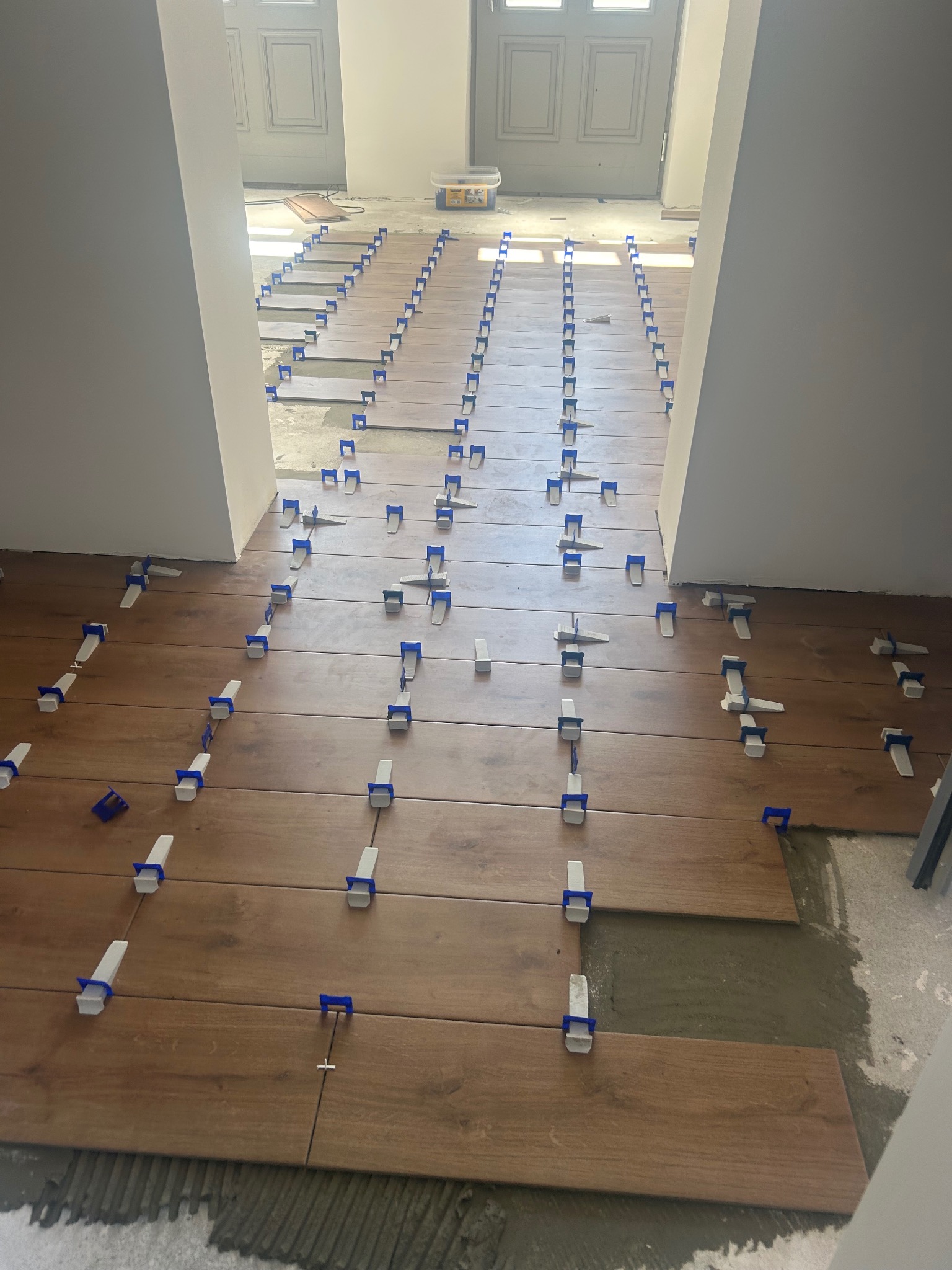 Układanie podłogi z płytek imitujących drewno, z użyciem systemów poziomujących z niebieskimi klinami, widoczne świeże zaprawy i przygotowane podłoże.