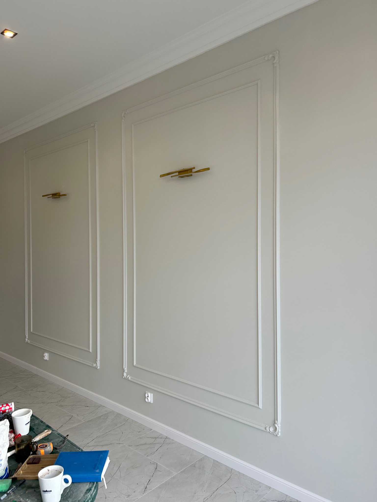 Ściana w jasnych odcieniach szarości z białymi, dekoracyjnymi panelami ściennymi i złotymi kinkietami, widoczne narzędzia malarskie na stole.