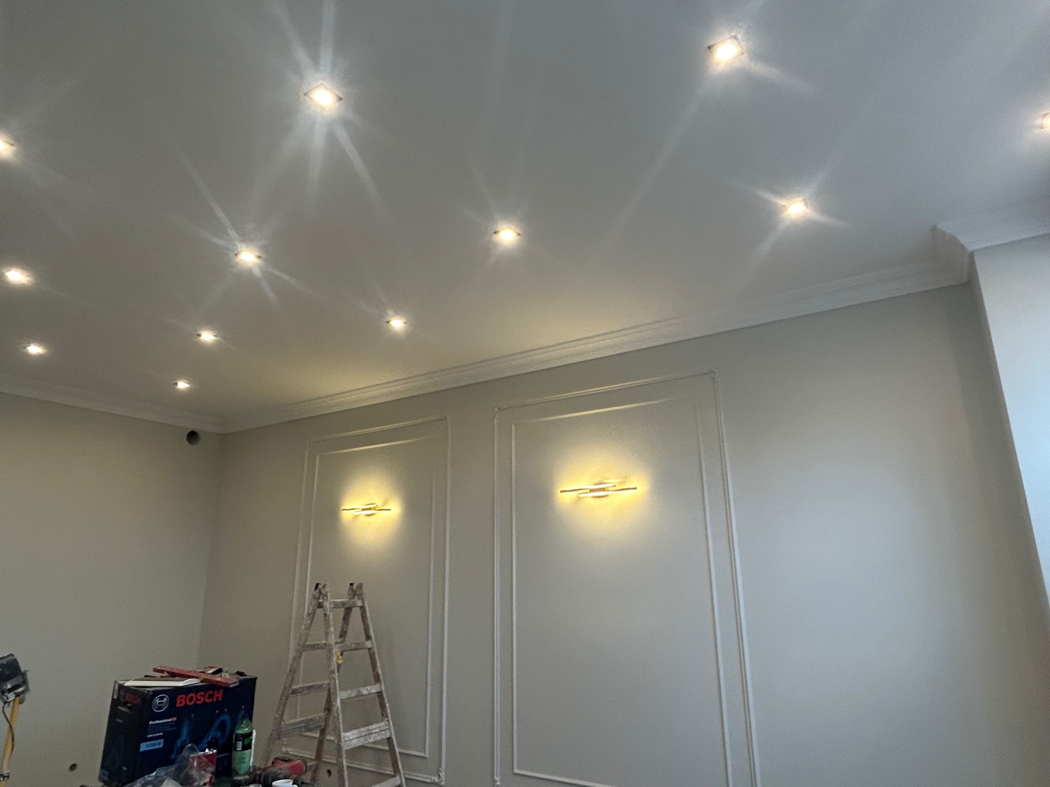 Wnętrze pokoju z nowymi punktowymi lampami sufitowymi i dwoma ozdobnymi lampami ściennymi, drabina oparta o ścianę, narzędzia i pudełko Bosch na podłodze.