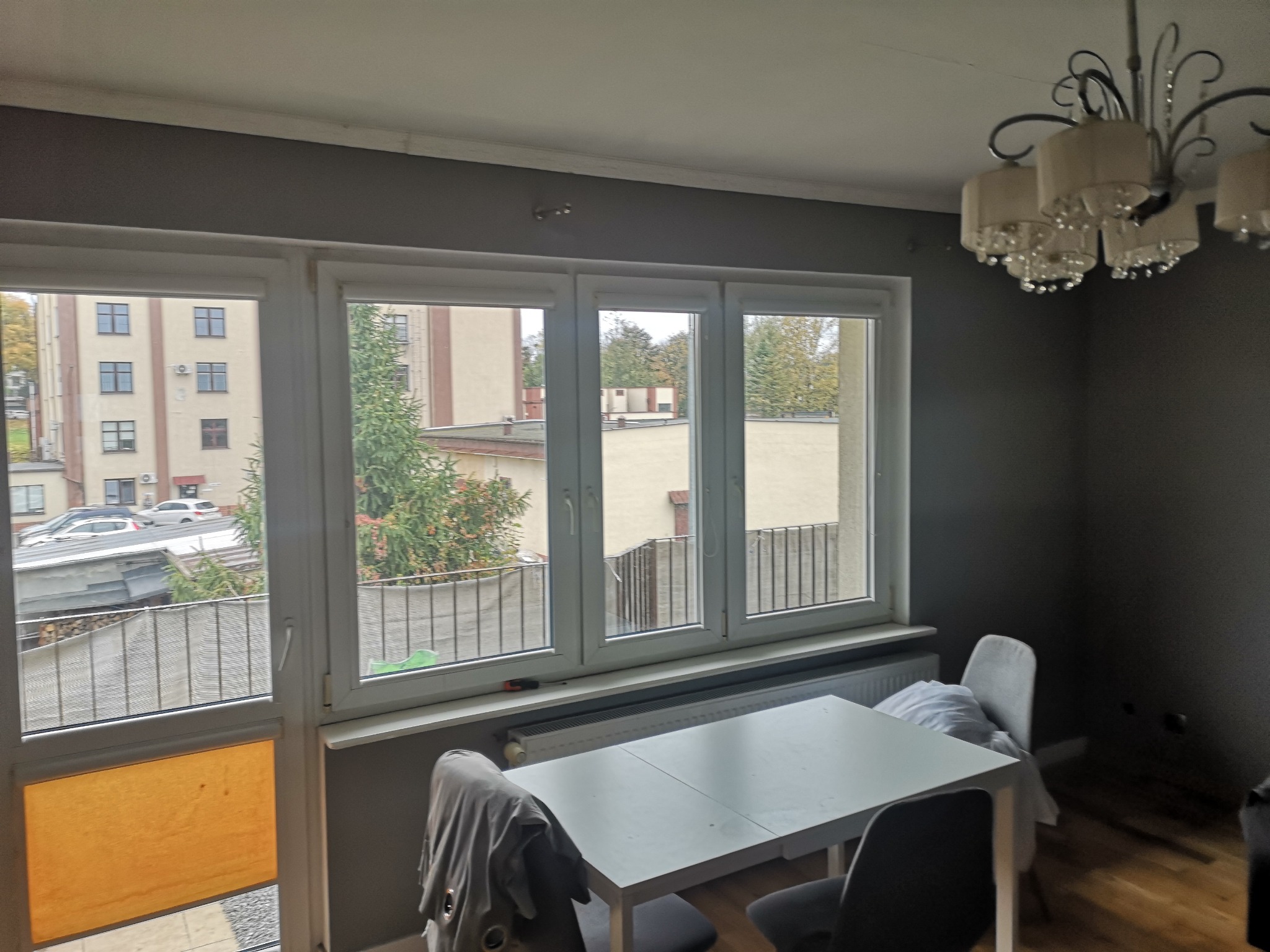 Wnętrze pokoju z dużym oknem podzielonym na trzy części, przez które widać budynek mieszkalny i drzewa. Na pierwszym planie biały stół z krzesłami i ozdobny żyrandol u góry po prawej stronie.